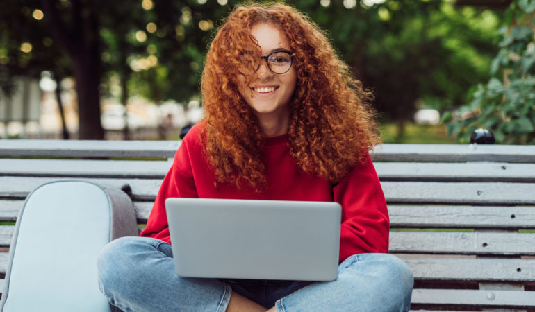 Happy red haired student with laptop sitting on bench